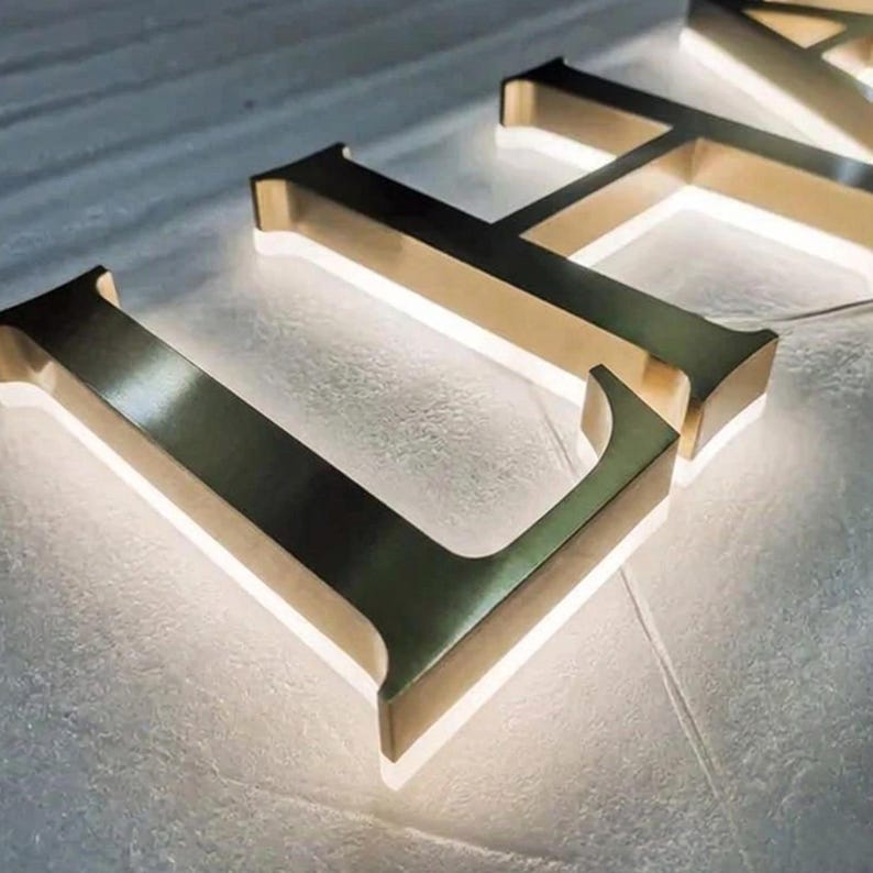 Puede incluir: Un conjunto de letras de metal en tono dorado tridimensionales con una tipograf&iacute;a serif. Cada letra tiene una superficie lisa y reflectante, iluminada desde atr&aacute;s con un suave resplandor blanco que crea un efecto de halo.