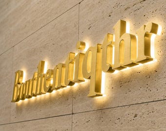Puede incluir: Un letrero de metal dorado en tres dimensiones que muestra la palabra "Bouthentiqthr" en una fuente con serifa. Las letras están montadas sobre una pared de piedra beige texturizada y están iluminadas desde atrás, creando un halo brillante alrededor de cada carácter.