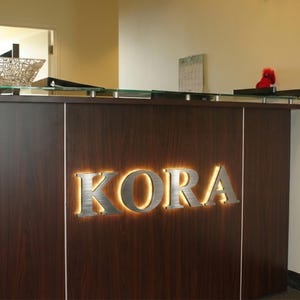 Puede incluir: Un mostrador de recepci&oacute;n de madera oscura con un letrero de metal cepillado con la palabra KORA en letras may&uacute;sculas. El letrero est&aacute; retroiluminado con un brillo c&aacute;lido, creando un efecto de halo sobre el panel de madera oscura del mostrador.