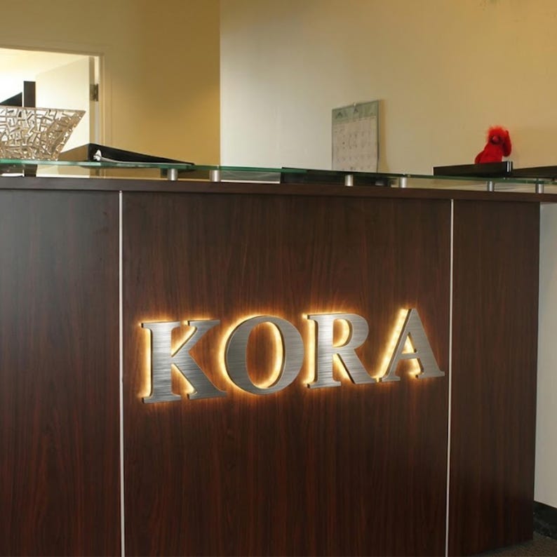 Puede incluir: Un mostrador de recepci&oacute;n de madera oscura con un letrero de metal cepillado con la palabra KORA en letras may&uacute;sculas. El letrero est&aacute; retroiluminado con un brillo c&aacute;lido, creando un efecto de halo sobre el panel de madera oscura del mostrador.