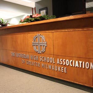 Puede incluir: Un mostrador de recepci&oacute;n de madera con un emblema de cruz plateada y el texto THE LUTHERAN HIGH SCHOOL ASSOCIATION OF GREATER MILWAUKEE en letras plateadas en relieve. El mostrador tiene un acabado de madera clara y una estructura rectangular.