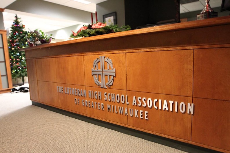 Puede incluir: Un mostrador de recepci&oacute;n de madera con un emblema de cruz plateada y el texto THE LUTHERAN HIGH SCHOOL ASSOCIATION OF GREATER MILWAUKEE en letras plateadas en relieve. El mostrador tiene un acabado de madera clara y una estructura rectangular.