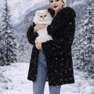 May include: A woman wearing a black coat, light blue jeans, and a cream-colored beanie holds a fluffy white cat in a snowy winter scene. The cat has light-colored fur and is being held close to the woman's chest.