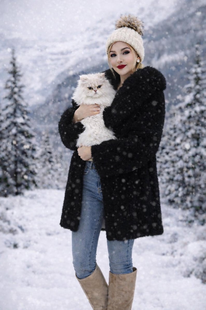 May include: A woman wearing a black coat, light blue jeans, and a cream-colored beanie holds a fluffy white cat in a snowy winter scene. The cat has light-colored fur and is being held close to the woman's chest.