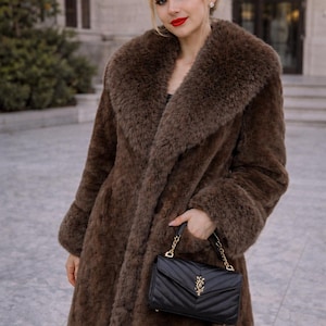 May include: A long, brown faux fur coat with a large collar and cuffs. The coat is worn with a black, quilted handbag with a gold chain strap. The woman is wearing red lipstick.