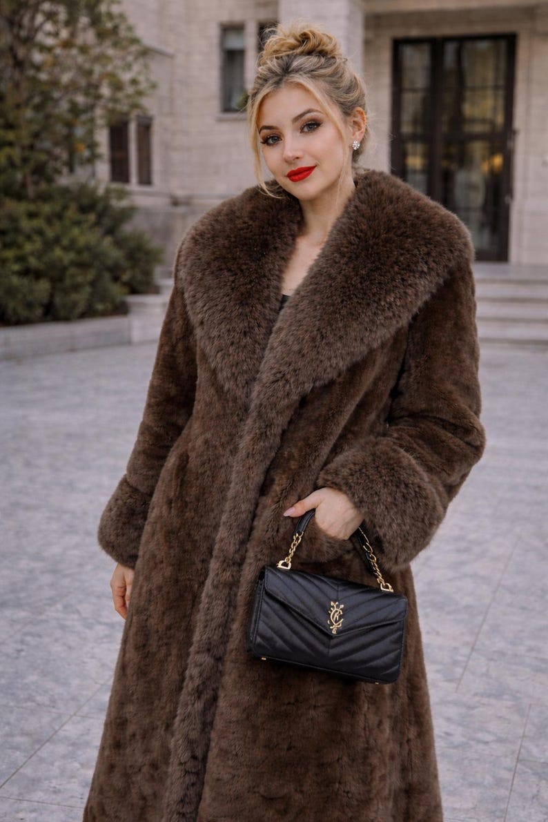 May include: A long, brown faux fur coat with a large collar and cuffs. The coat is worn with a black, quilted handbag with a gold chain strap. The woman is wearing red lipstick.