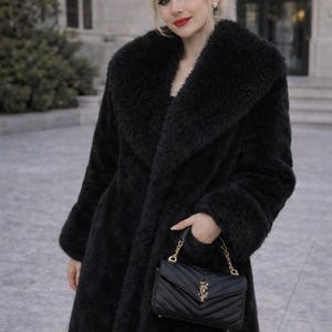 May include: A long, black faux fur coat with a wide collar and cuffs. A black, quilted leather handbag with a gold chain strap is held in the hand. The coat is worn outdoors.