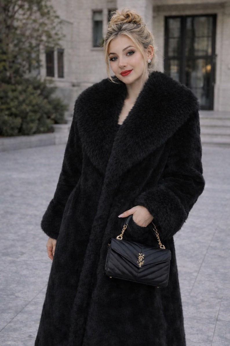 May include: A long, black faux fur coat with a wide collar and cuffs. A black, quilted leather handbag with a gold chain strap is held in the hand. The coat is worn outdoors.
