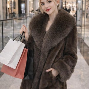 May include: A long, brown faux fur coat with a large collar and pockets. The coat is worn by a person holding shopping bags in white, coral, and black. The background shows a shopping mall.
