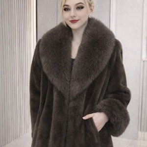 May include: A long, brown faux fur coat with a wide collar and cuffs. The coat has a classic design with a tailored fit and a pocket. The coat is a luxurious outerwear piece, perfect for cold weather.