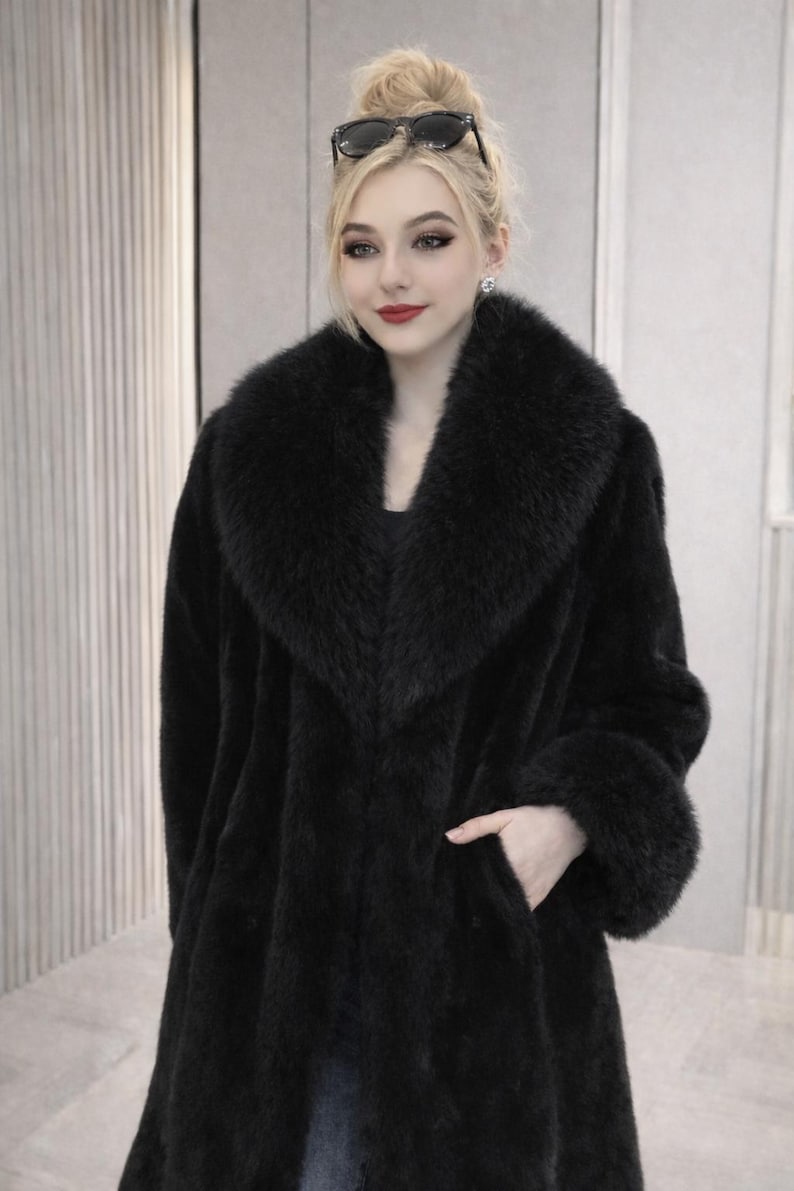 May include: A long, black faux fur coat with a wide collar and cuffs. The coat has a classic design with a tailored fit and a single pocket. The model is wearing sunglasses on top of their head.