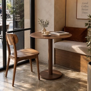 May include: A round brown wooden pedestal table paired with a matching wooden chair. On the table sits a small white ceramic vase with green sprigs, a white coffee cup on a saucer, and a closed book. The furniture has a smooth, matte finish.