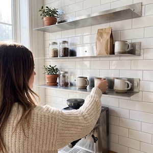 Puede incluir: Tres estantes horizontales de metal cepillado montados en una pared de azulejos blancos. Los estantes sostienen dos plantas en macetas de terracota, tres frascos de vidrio con contenido, una bolsa de papel marrón y cuatro tazas de cerámica.