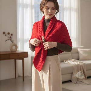 Puede incluir: Un chal rojo vivo de punto trenzado con un cierre decorativo. El chal está drapeado sobre los hombros y abrochado en la parte delantera. El cierre es de color marrón claro. El chal es un accesorio cálido.