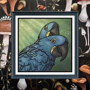 May include: A square framed cross-stitch artwork featuring two blue hyacinth macaws with yellow eye rings and dark beaks. The birds are positioned against a textured green background with light rays, all enclosed in a simple black frame.