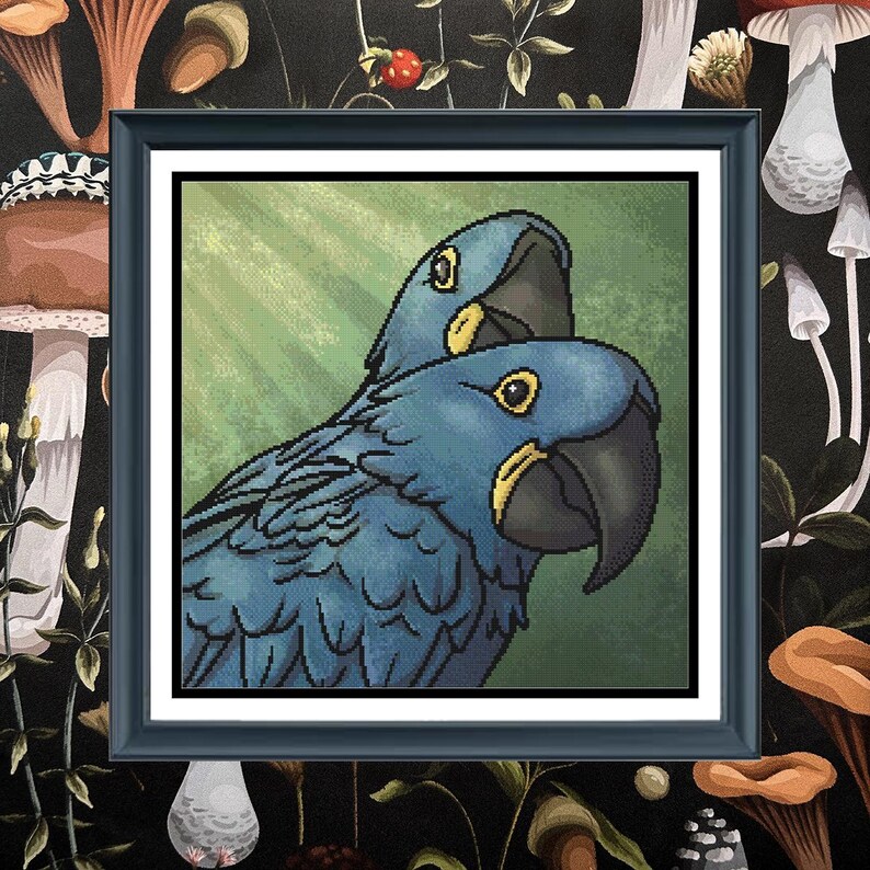 May include: A square framed cross-stitch artwork featuring two blue hyacinth macaws with yellow eye rings and dark beaks. The birds are positioned against a textured green background with light rays, all enclosed in a simple black frame.