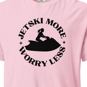 以下が含まれることがあります： 薄いピンク色のTシャツ。黒いグラフィックは、ジェットスキーに乗る人物のシルエットと、周りに「Jet Ski More Worry Less」という言葉が円形に配置されています。クルーネックと半袖のデザインです。