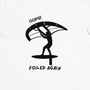 May include: White t-shirt with a black graphic of a person windsurfing, with the text "OOPS!" above the figure and "FOILED AGAIN" below. The shirt has a crew neck and short sleeves.