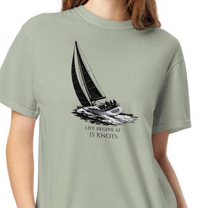 May include: Sage green t-shirt featuring a black and white illustration of a sailboat on the water. The text "LIFE BEGINS AT 15 KNOTS" is printed below the image. The shirt is made of a soft material.