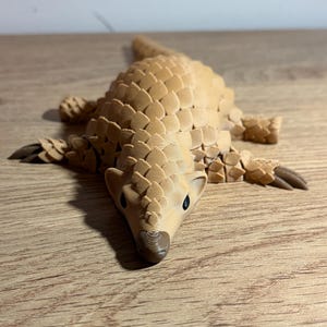 May include: A tan 3D-printed pangolin figurine. The pangolin is lying down with its legs spread. The body is covered in overlapping scales, and the head has small black eyes and a brown snout. The figurine is on a wooden surface.