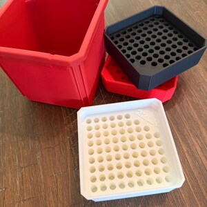 May include: A selection of plastic containers and trays in red, black, and white. The red container is a large, open-top box. The black tray has a grid of holes. The white tray also has a grid of holes. The items are on a wooden surface.