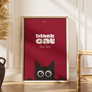 Peut inclure: Une affiche encadrée avec un fond rouge et les mots "black cat lives here" en blanc. Une illustration de chat noir apparaît en bas. L'affiche est dans un cadre doré, appuyée contre un mur blanc.