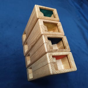 Könnte beinhalten: Vier gestapelte Holzkisten mit offenen Oberseiten, die jeweils einen unterschiedlich farbigen Innenraum freilegen: grün, gelb, blau und rot. Die Kisten sind aus hellem Holz gefertigt und haben eine rechteckige Form. Die Kisten sind übereinander gestapelt.