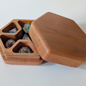 May include: A wooden, hexagonal dice box with a hinged lid, open to reveal several multi-sided dice. The box is made of a light brown wood and has compartments to hold the dice. The lid is open, resting beside the box.
