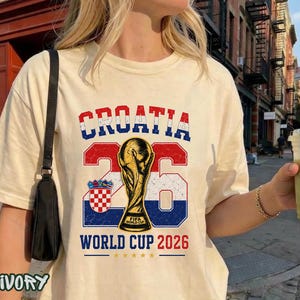 May include: An ivory t-shirt featuring a graphic with the text CROATIA in red, white, and blue, the number 26, a gold FIFA World Cup trophy, the Croatian coat of arms, and the text WORLD CUP 2026 with five small gold stars underneath the text.