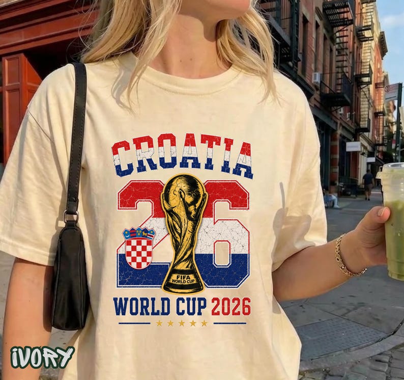 May include: An ivory t-shirt featuring a graphic with the text CROATIA in red, white, and blue, the number 26, a gold FIFA World Cup trophy, the Croatian coat of arms, and the text WORLD CUP 2026 with five small gold stars underneath the text.