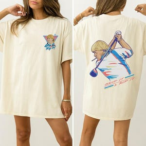 Könnte beinhalten: Cremefarbenes T-Shirt mit einer Grafik eines Golfspielers und dem Text "Grip It & Rip It". Die Vorderseite des Shirts hat eine kleinere Version der Grafik. Das Design enthält blaue, rosa und weiße Akzente.