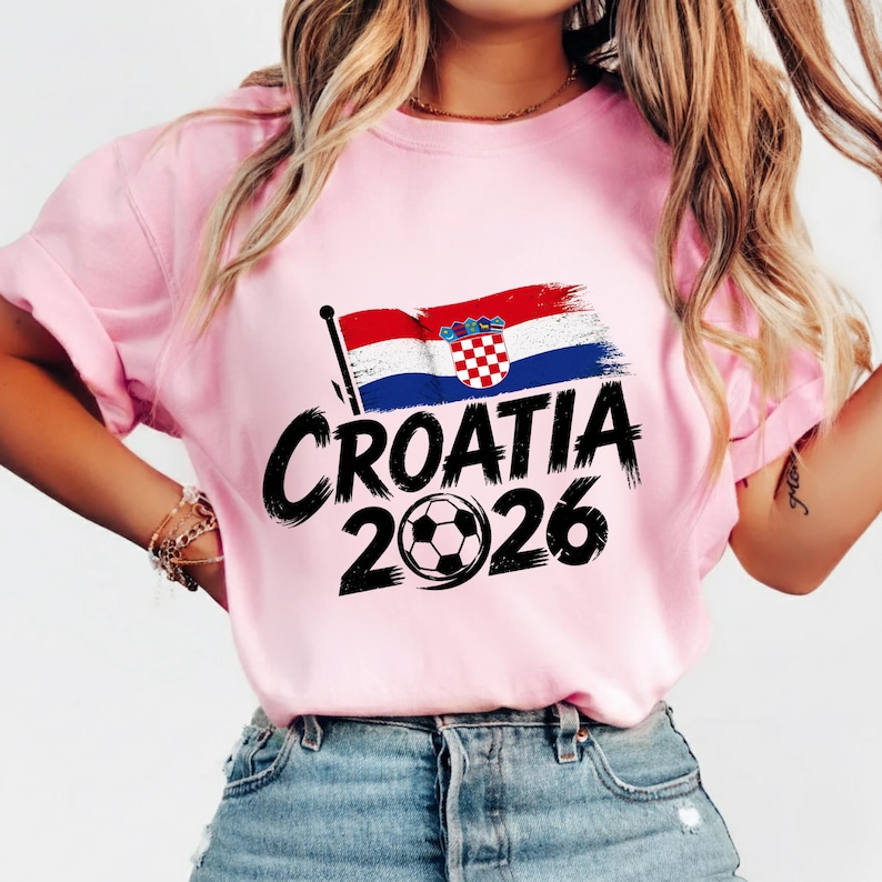Puede incluir: Una camiseta de manga corta de color rosa claro con un estampado gr&aacute;fico de la bandera de Croacia, el texto "CROATIA" en una fuente negra gruesa estilo pincelada y el a&ntilde;o "2026" con un bal&oacute;n de f&uacute;tbol sustituyendo al cero.