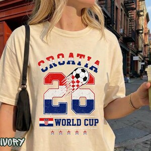 May include: An ivory t-shirt featuring a graphic with the text CROATIA 26 WORLD CUP. The design includes a soccer ball, the number 26 in red, white, and blue, a small Croatian flag, and five stars, all presented in a distressed, vintage-style print.