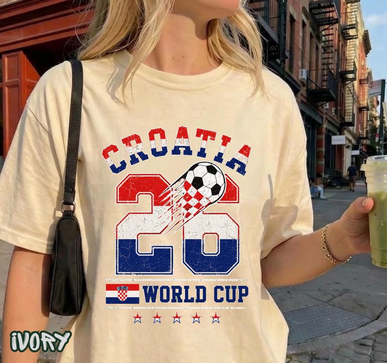 May include: An ivory t-shirt featuring a graphic with the text CROATIA 26 WORLD CUP. The design includes a soccer ball, the number 26 in red, white, and blue, a small Croatian flag, and five stars, all presented in a distressed, vintage-style print.
