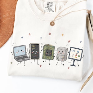 May include: An off-white t-shirt featuring a graphic of five smiling, anthropomorphic computer components including a laptop, a code notebook, a circuit board, a server tower, and a monitor displaying a flowchart, accented by small colorful stars.
