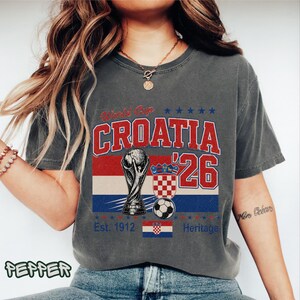 May include: A charcoal gray t-shirt featuring a graphic print with the text World Cup CROATIA '26. The design includes a trophy illustration, a soccer ball, the Croatian flag, and the text Est. 1912 Heritage in red, white, and blue colors.