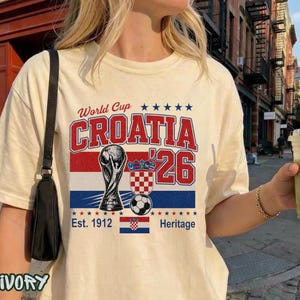 May include: An ivory t-shirt featuring a graphic print with the text World Cup CROATIA '26. The design includes a trophy illustration, a soccer ball, the Croatian coat of arms, a small Croatian flag, and the text Est. 1912 Heritage with blue stars.