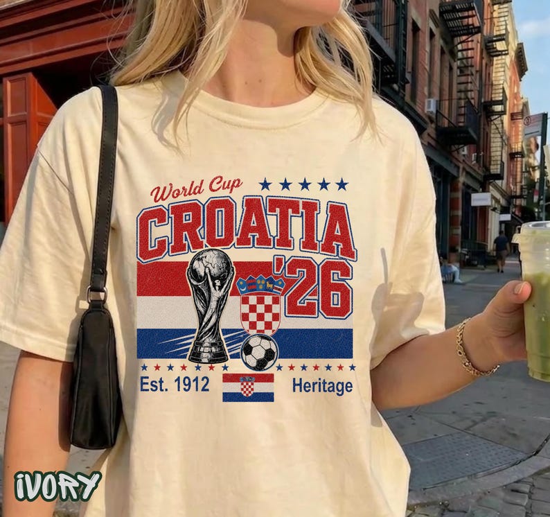 May include: An ivory t-shirt featuring a graphic print with the text World Cup CROATIA '26. The design includes a trophy illustration, a soccer ball, the Croatian coat of arms, a small Croatian flag, and the text Est. 1912 Heritage with blue stars.
