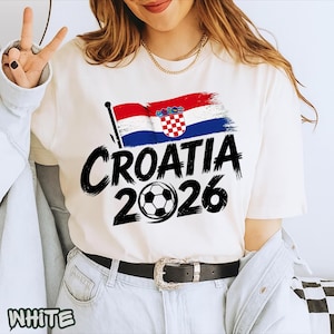Puede incluir: Una camiseta blanca de manga corta con un gr&aacute;fico de la bandera de Croacia, el texto "CROATIA" en una fuente negra gruesa estilo pincelada, y el a&ntilde;o "2026" con un bal&oacute;n de f&uacute;tbol reemplazando el cero, impreso en la parte delantera.