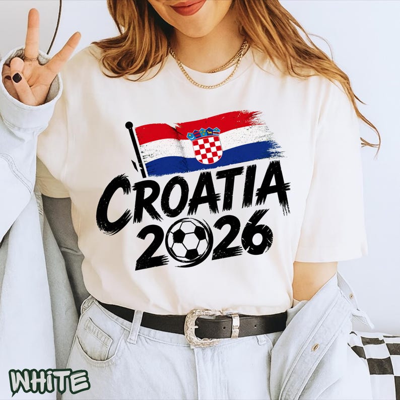 Puede incluir: Una camiseta blanca de manga corta con un gr&aacute;fico de la bandera de Croacia, el texto "CROATIA" en una fuente negra gruesa estilo pincelada, y el a&ntilde;o "2026" con un bal&oacute;n de f&uacute;tbol reemplazando el cero, impreso en la parte delantera.