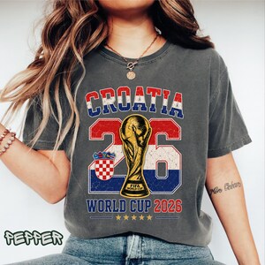 May include: A charcoal gray short-sleeve t-shirt featuring a graphic with the text CROATIA, the number 26, and WORLD CUP 2026. The design includes a gold FIFA World Cup trophy illustration, the Croatian coat of arms, and five gold stars.