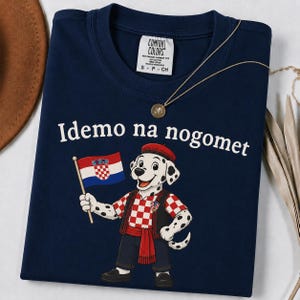 Op de afbeelding: Een marineblauw t-shirt met een cartoon dalmati&euml;r die een rode baret en een rood-wit geblokt vest draagt. De hond houdt een Kroatische vlag vast en de tekst "Idemo na nogomet" is in het wit boven de afbeelding gedrukt. Comfort Colors label.