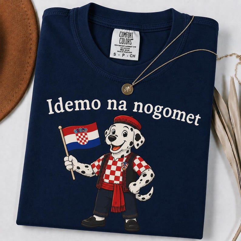 Op de afbeelding: Een marineblauw t-shirt met een cartoon dalmati&euml;r die een rode baret en een rood-wit geblokt vest draagt. De hond houdt een Kroatische vlag vast en de tekst "Idemo na nogomet" is in het wit boven de afbeelding gedrukt. Comfort Colors label.