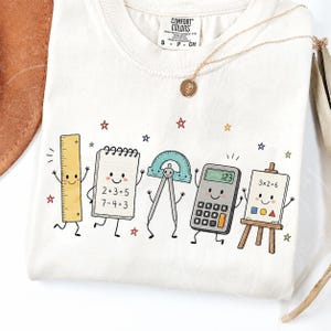 Op de afbeelding: Een gebroken wit t-shirt met een afbeelding van vijf antropomorfe schoolspullen: een liniaal, een spiraalblok met sommen, een geodriehoek, een rekenmachine en een kleine ezel met vormen, omringd door kleurrijke sterren.