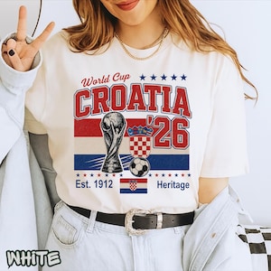 May include: A white short-sleeve t-shirt featuring a graphic with the text World Cup CROATIA '26. The design includes a soccer trophy, a soccer ball, the Croatian coat of arms, a small Croatian flag, and the text Est. 1912 Heritage in red and blue.