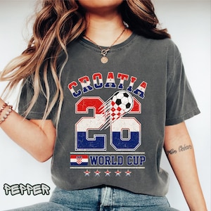 May include: A charcoal gray t-shirt featuring a distressed graphic with the text CROATIA 26 WORLD CUP. The design includes a soccer ball, the Croatian flag, and five stars. The number 26 is large, filled with the red, white, and blue flag pattern.