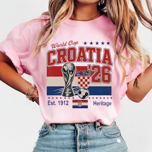 May include: A light pink t-shirt featuring a graphic with the text World Cup CROATIA '26. The design includes a soccer trophy, a soccer ball, the Croatian coat of arms, a small Croatian flag, and the text Est. 1912 Heritage with red and blue stars.