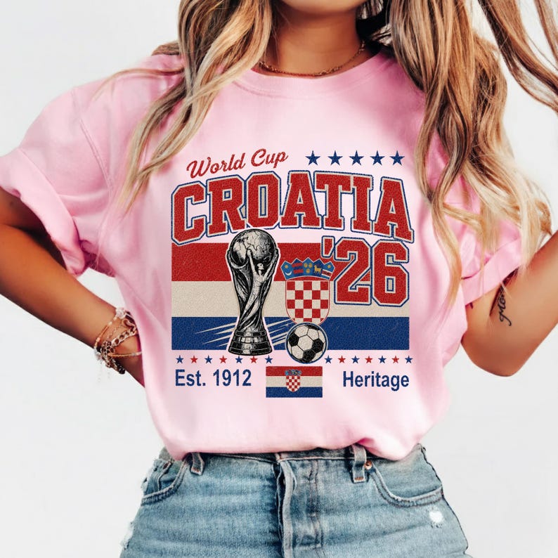May include: A light pink t-shirt featuring a graphic with the text World Cup CROATIA '26. The design includes a soccer trophy, a soccer ball, the Croatian coat of arms, a small Croatian flag, and the text Est. 1912 Heritage with red and blue stars.