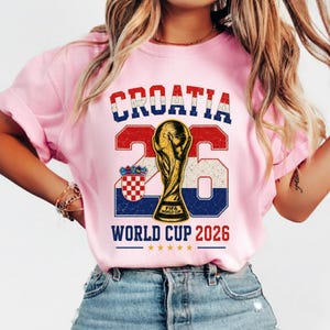 May include: A light pink t-shirt featuring a graphic with the text CROATIA in red, white, and blue, the number 26, a gold FIFA World Cup trophy, the Croatian coat of arms, and the text WORLD CUP 2026 with five small gold stars underneath.