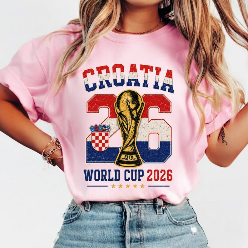 May include: A light pink t-shirt featuring a graphic with the text CROATIA in red, white, and blue, the number 26, a gold FIFA World Cup trophy, the Croatian coat of arms, and the text WORLD CUP 2026 with five small gold stars underneath.